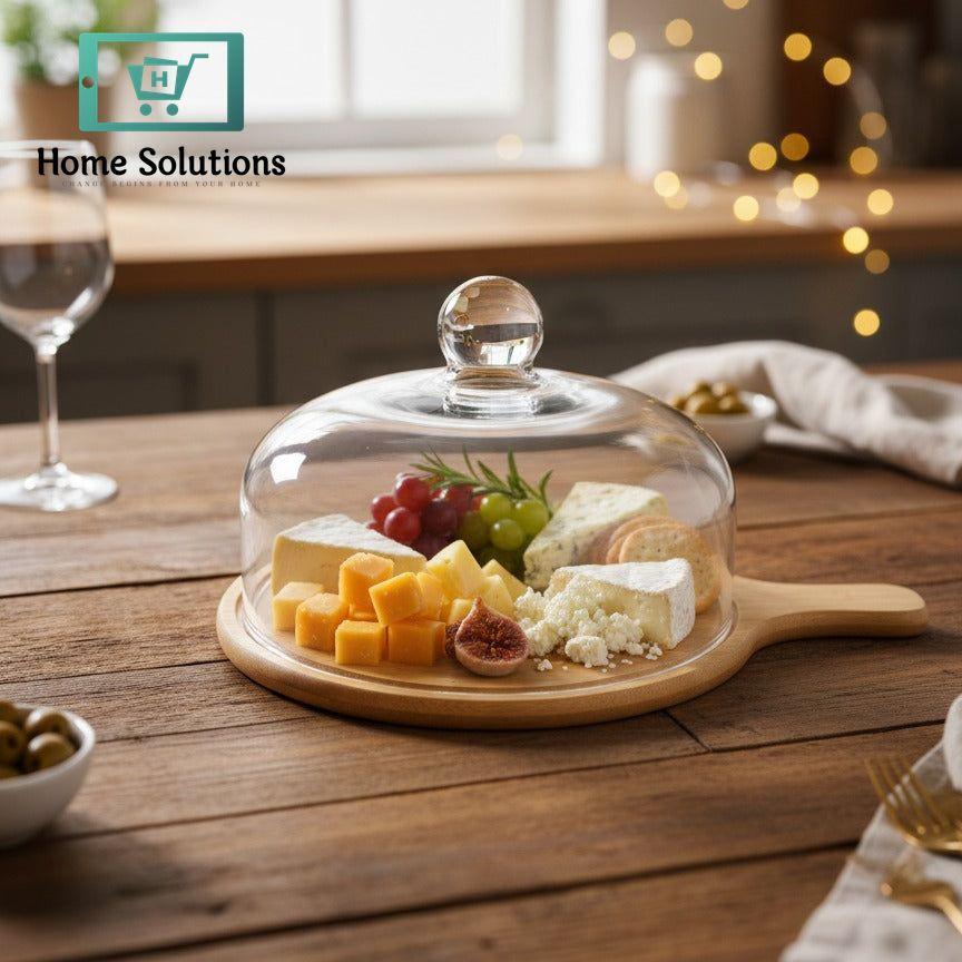 Glass dome on a wooden board with assorted cheeses and fruits on a wooden table.
