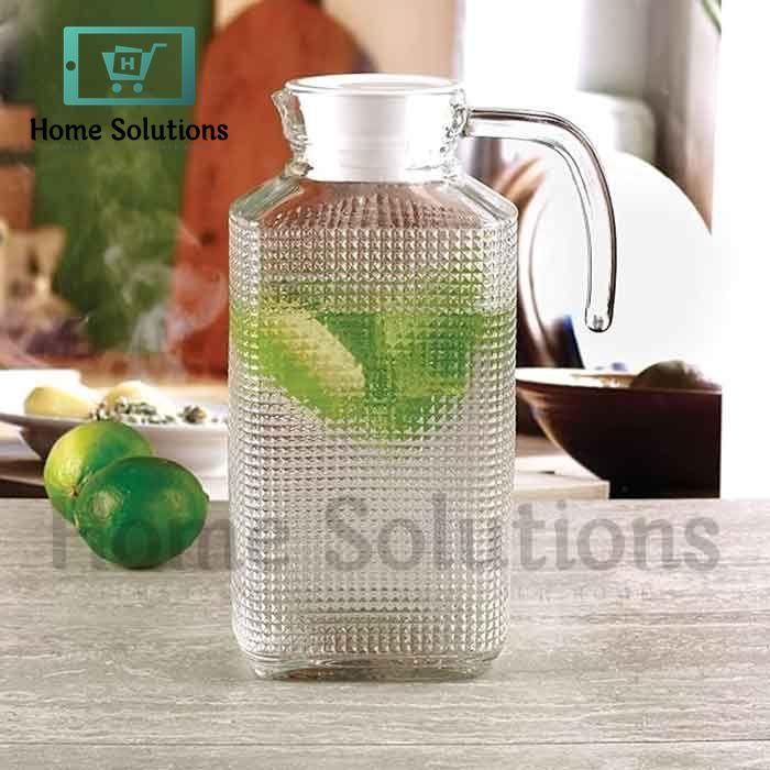 Clear glass pitcher with textured surface on a table with limes and a blurred background.