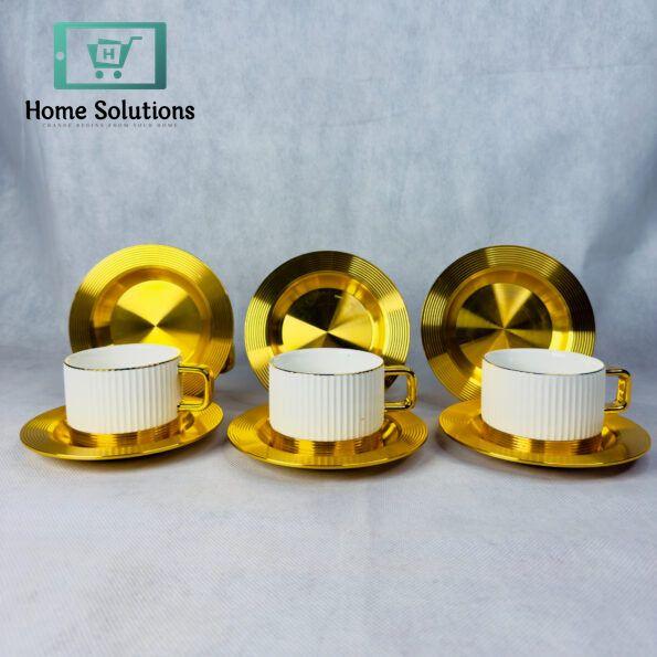 Set of 6 elegant ribbed ceramic coffee and tea cups with gold metal bases and matching gold metal saucers, stacked on a marble surface with blurred golden background