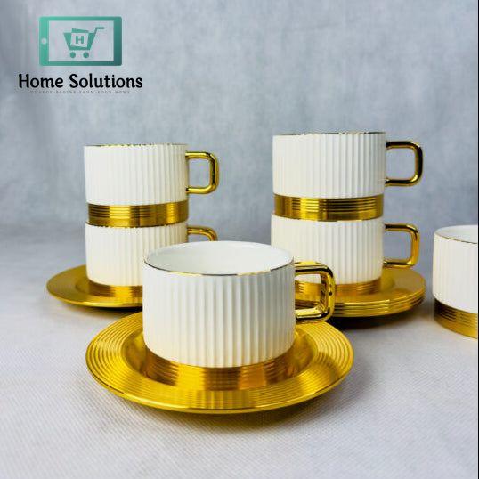 Set of 6 elegant ribbed ceramic coffee and tea cups with gold metal bases and matching gold metal saucers, stacked on a marble surface with blurred golden background