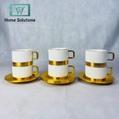 Set of 6 elegant ribbed ceramic coffee and tea cups with gold metal bases and matching gold metal saucers, stacked on a marble surface with blurred golden background